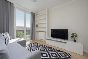 Ein modernes Wohnzimmer mit einer Couch, einem Fernseher auf einem weißen Schrank, einem gemusterten Teppich und einem Balkon mit Vorhängen.