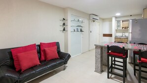 A living room with a black leather sofa, red cushions, a dining area with a table and chairs, and a kitchen in the background.