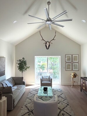 Ein geräumiges Wohnzimmer mit hoher Decke, Deckenventilator, einem aufgehängten Hirschschädel, einem großen Fenster, einer Couch, einem Couchtisch und einem Blumentopf.