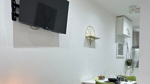 Ein modernes Wohnzimmer mit Flachbildfernseher, einem kleinen Tisch mit Lampe und einem Essbereich mit grünen Stühlen.