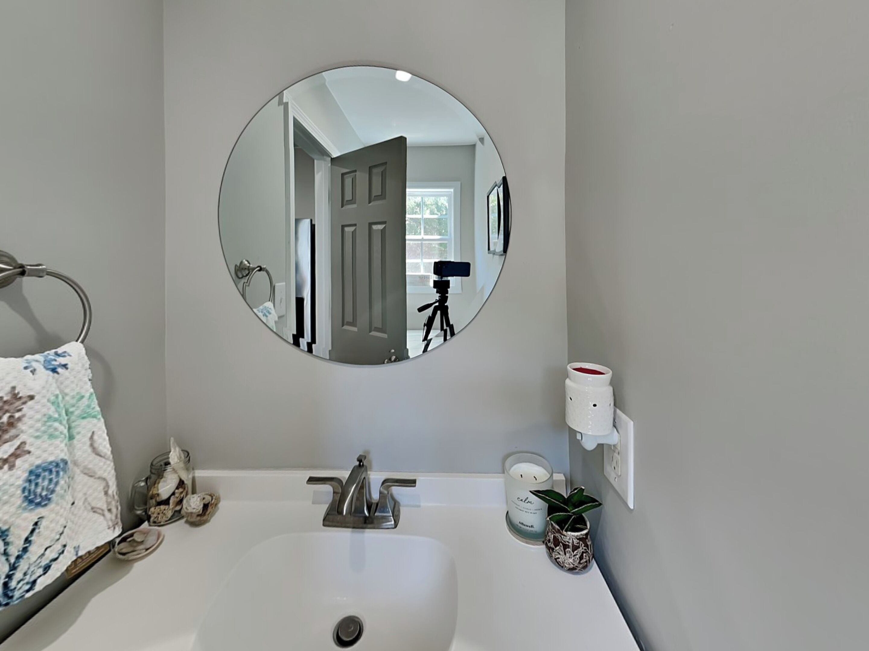 A bathroom with a round mirror, a sink, a towel rack, a candle, and a small plant.