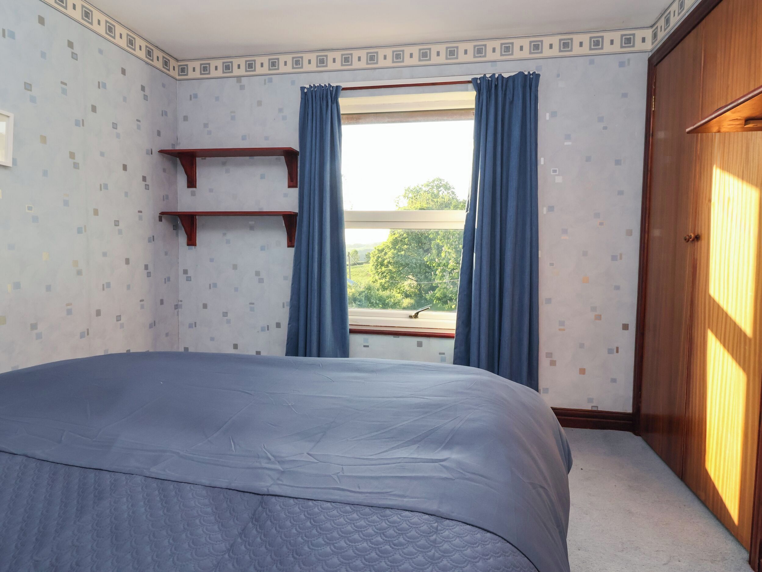 A bedroom with a bed, a window with curtains, a wooden wardrobe, and a shelf.