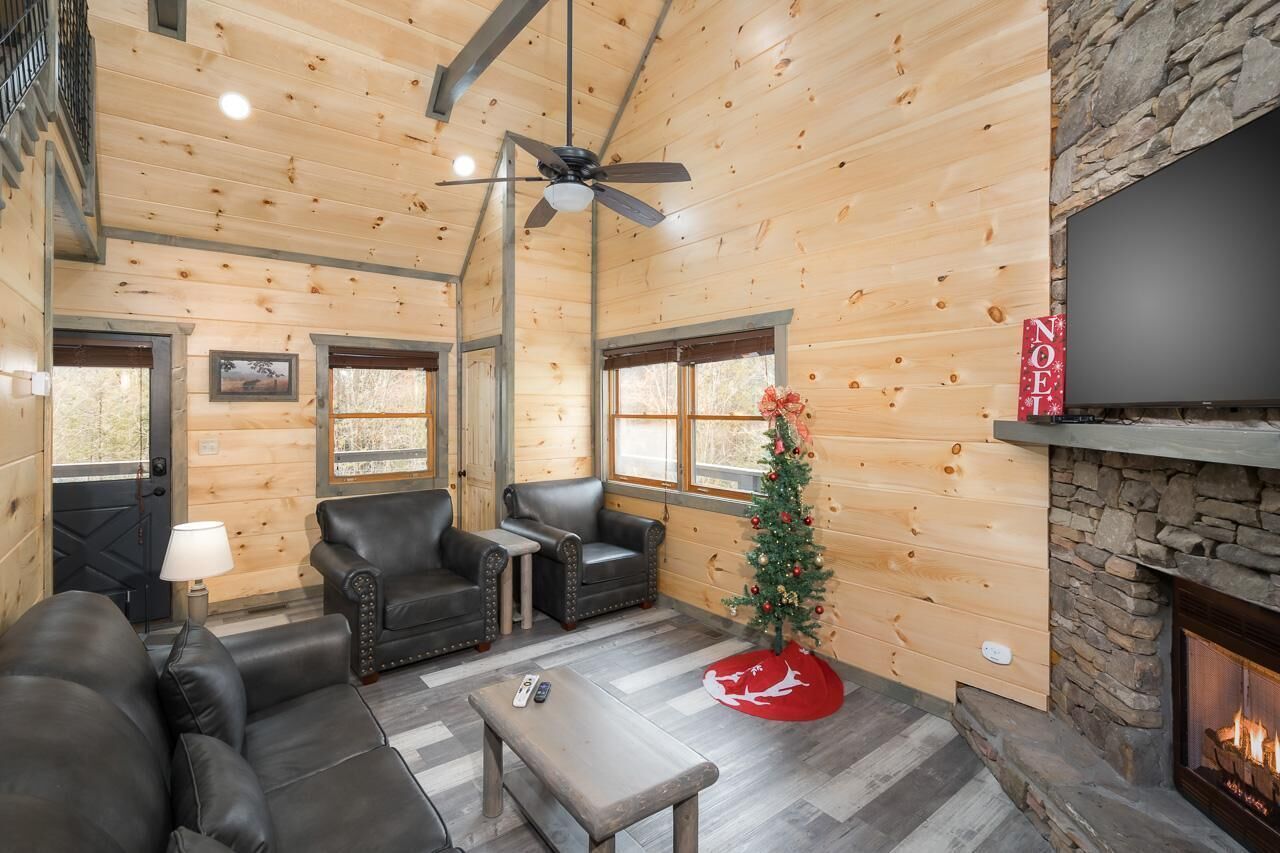 A cozy cabin interior with a fireplace, leather seating, a small Christmas tree, and a ceiling fan.