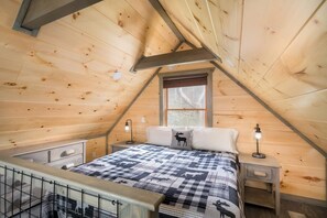 Un dormitorio con techo inclinado, una cama con colcha a cuadros, dos lámparas de noche, un ventanal y una mesita de noche pequeña.