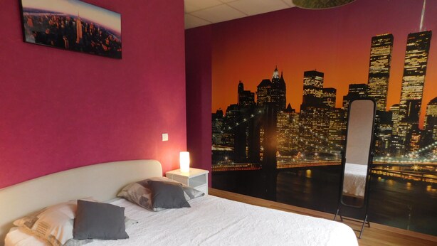 Una habitación de hotel con cama, almohadas, una mesita de noche, un cuadro en la pared y un mural con vista a la ciudad.