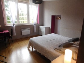 A bedroom with a bed, bedside table, a desk, a chair, a radiator, and a window with curtains.