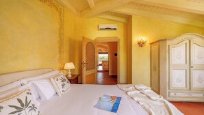 A hotel room with a bed, a nightstand, an air conditioning unit, a wardrobe, and a hallway leading to an outdoor area. - NG Collection Cala del Faro 206 (Arzachena)