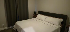A neatly made bed with a white bedspread and pillows, a wooden nightstand with a lamp, and a dark-colored headboard.