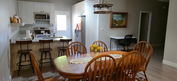 A modern kitchen with white cabinets, a microwave, and a refrigerator. There is a dining area with wooden chairs and a table, and a painting on the wall.
