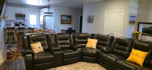 A living room with a sectional sofa, a dining area with chairs, a kitchen with appliances, and artwork on the walls.