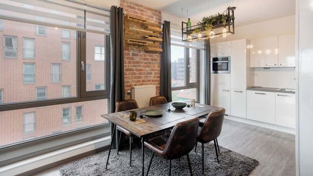 Une cuisine moderne avec des meubles blancs, un mur d’accent en briques et un coin repas comprenant une table et des chaises en bois.