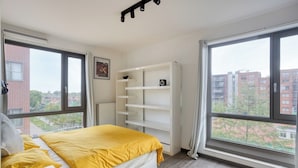Une chambre avec un lit, une étagère blanche et de grandes fenêtres donnant sur la ville.