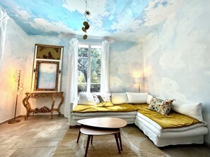 A room with a ceiling painted to resemble clouds, a white sofa with yellow and floral cushions, a round wooden table, and a large window with curtains. - Villa le Verger in Aix-en-Provence (Aix-en-Provence)