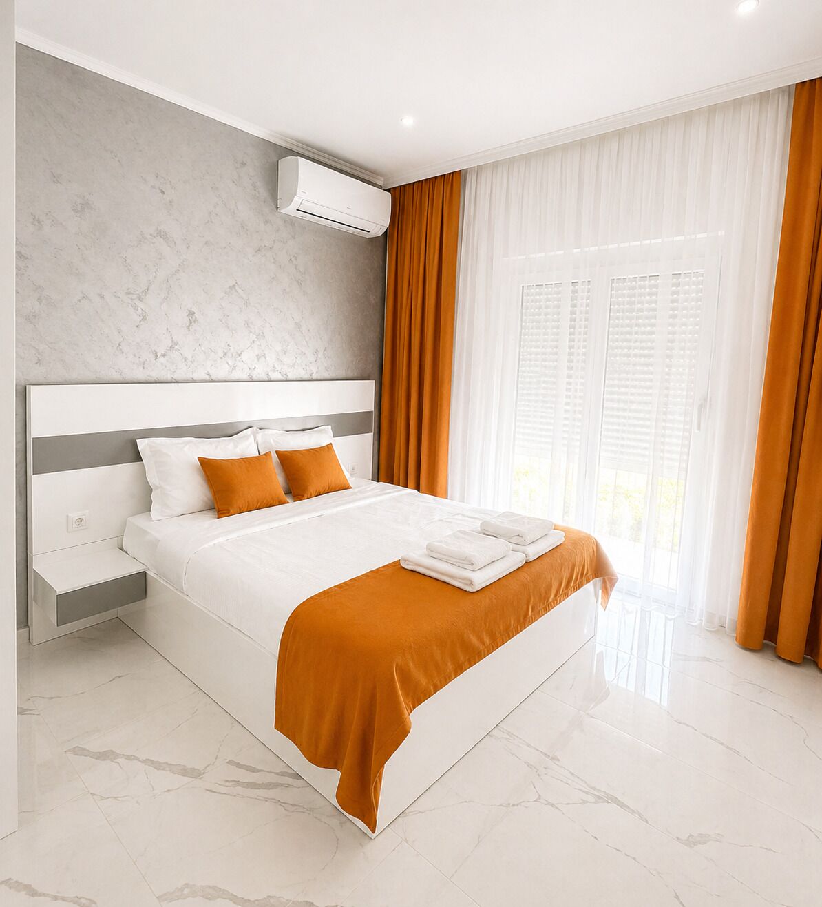 A modern hotel room with a marble floor, a large bed with white bedding and orange accents, a built-in air conditioning unit, and large windows with curtains.
