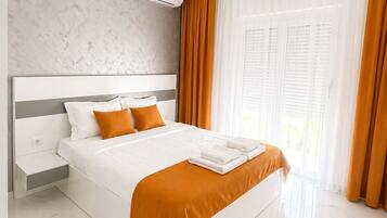 A modern hotel room with a marble floor, a large bed with white bedding and orange accents, a built-in air conditioning unit, and large windows with curtains.