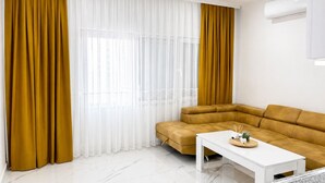 Een moderne woonkamer met een hoekbank, een witte salontafel en een groot raam met transparante witte gordijnen en mosterdgele gordijnen.