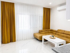 Ein modernes Wohnzimmer mit einem Ecksofa, einem weißen Couchtisch und einem großen Fenster mit transparenten weißen und senffarbenen Vorhängen.