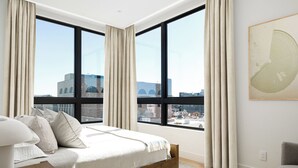 A bedroom with a large bed, a view of the city through large windows, and a painting on the wall. - Downtown Traverse City Penthouse w/ Private Rooftop (Traverse City)