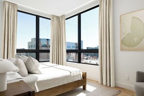 A bedroom with a large bed, a view of the city through large windows, and a painting on the wall. - Downtown Traverse City Penthouse w/ Private Rooftop (Traverse City)