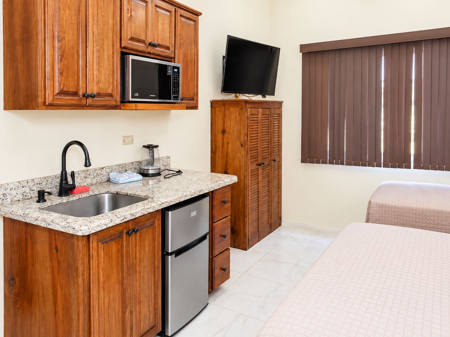 A compact kitchen with a microwave, sink, and refrigerator, a television mounted on the wall, and a bed with a window.