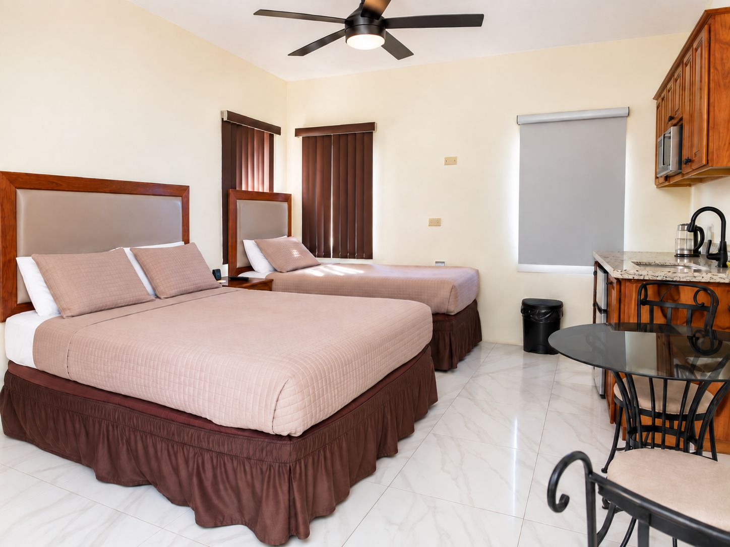 A hotel room with two beds, a ceiling fan, a kitchenette, and a dining area.