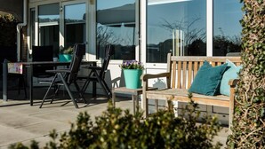 Un patio al aire libre con una mesa y sillas, un banco de madera con cojines y plantas en macetas.