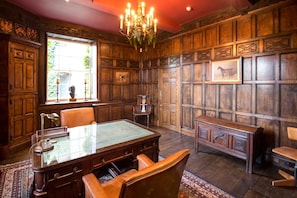 Une pièce avec un mur lambrissé en bois, un lustre, un bureau avec des fauteuils en cuir et une fenêtre donnant sur un paysage verdoyant.