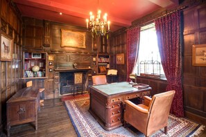 Une piÚce avec un bureau en bois, un fauteuil en cuir, des étagÚres, une cheminée et un lustre.