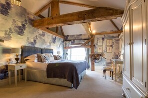 Une chambre Ă coucher avec un grand lit, des poutres en bois, une commode, une coiffeuse et une fenĂȘtre avec des rideaux.