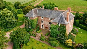 A large, historic stone house with a symmetrical facade, a central entrance, and a well-maintained garden with hedges and a lawn.