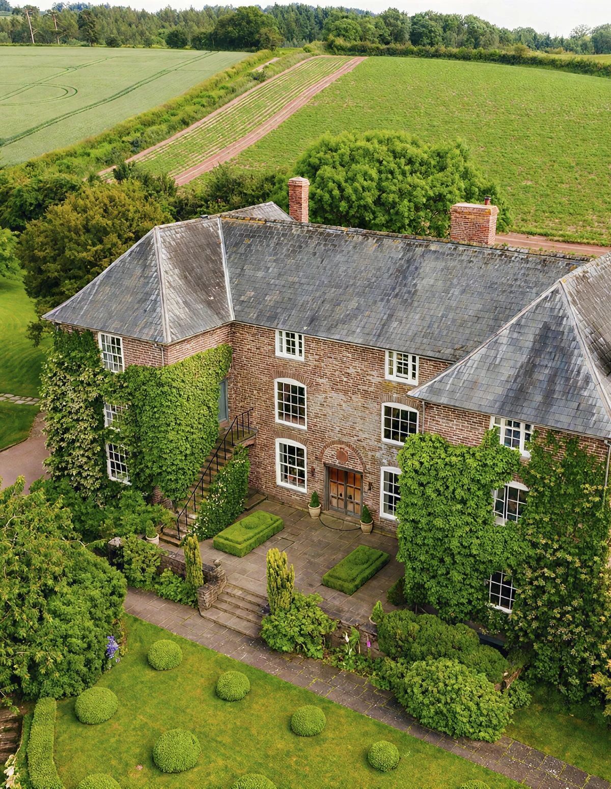 A large brick house with a steep roof, surrounded by a well-maintained garden and fields.