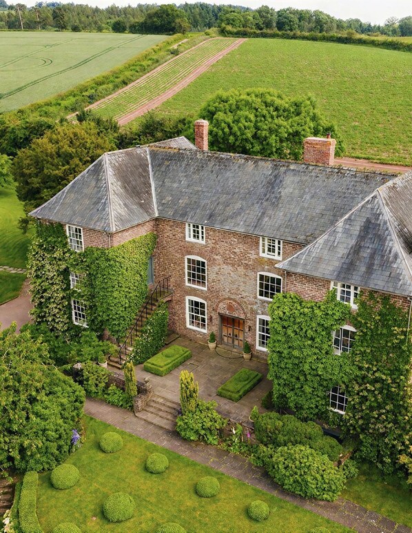 A large brick house with a steep roof, surrounded by a well-maintained garden and fields.
