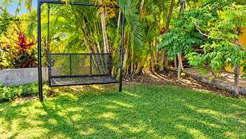 A black metal swing set in a lush garden with various tropical plants and a concrete wall.