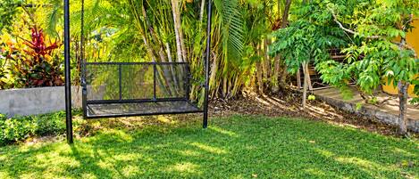 A black metal swing set in a lush garden with various tropical plants and a concrete wall.