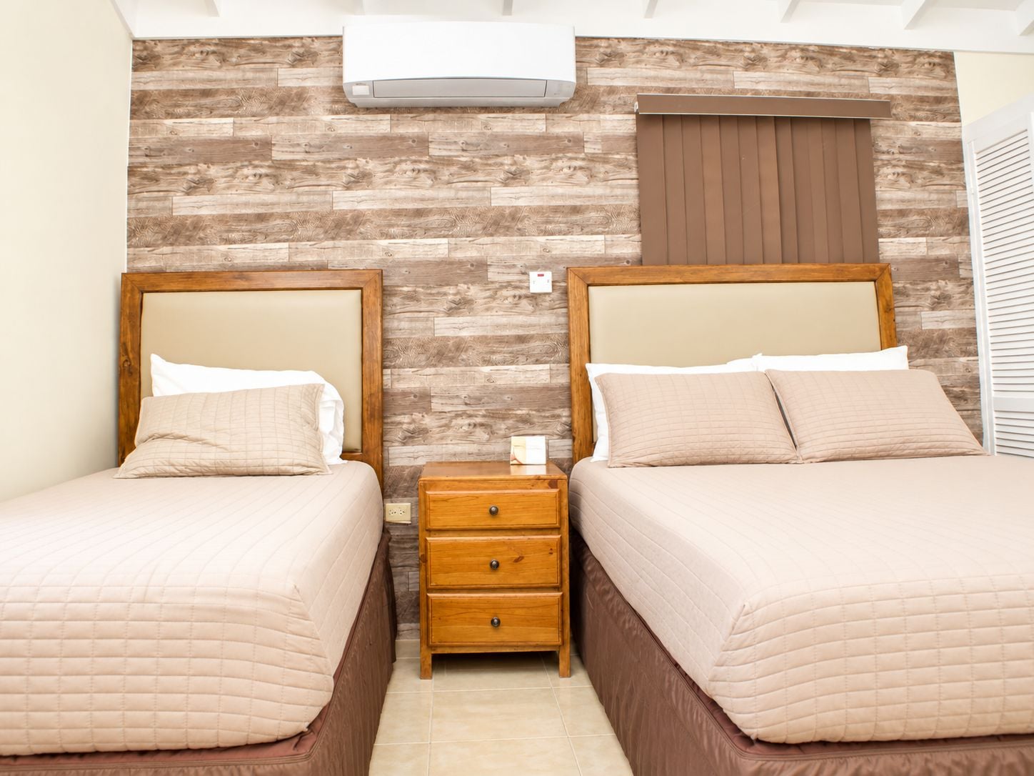 A hotel room with two beds, a wooden wall, a nightstand, and a window with blinds.