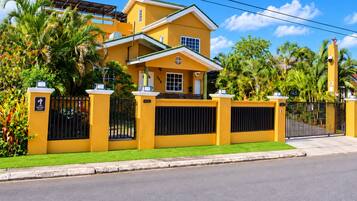 A yellow two-story house with a gated entrance, surrounded by lush greenery and palm trees.