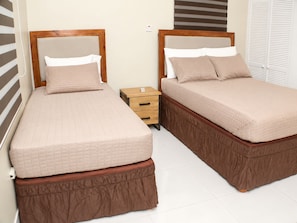Two twin beds with wooden frames and matching brown bed skirts, a small wooden nightstand with a lamp, and a white closet with sliding doors.