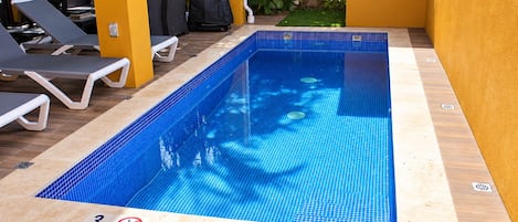 Una piscina rectangular interior con azulejos azules, rodeada de sillas de descanso y una pared amarilla.