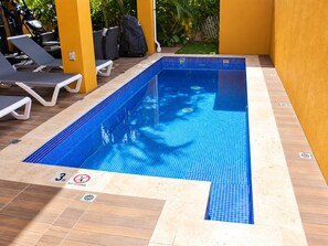 A rectangular indoor swimming pool with blue tiles, surrounded by lounge chairs and a yellow wall.