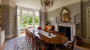 A dining room with a large wooden table, chairs, a chandelier, a fireplace, and a large window with a view of greenery. - Lakestone House - Five Bedroom Villa, Sleeps 10 (Windermere)