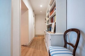 Ein schmaler Flur mit Holzboden, einem weißen Schrank mit Büchern und einem Holzstuhl mit Streifenpolster.
