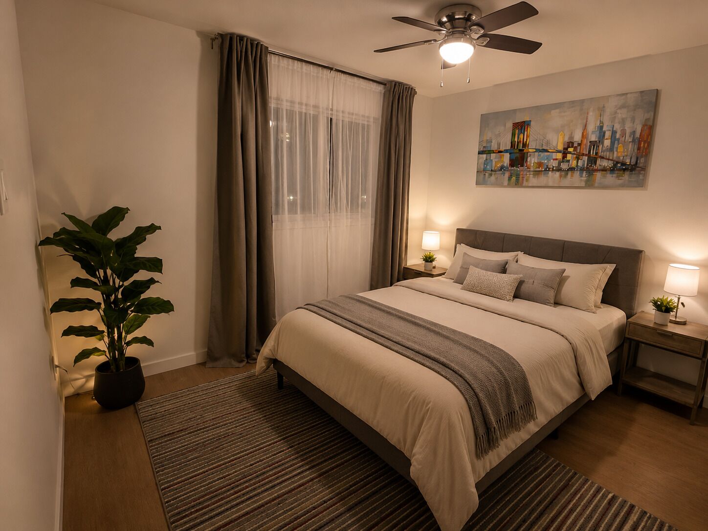 A well-lit bedroom with a bed, a ceiling fan, a nightstand with a lamp, a potted plant, and a painting on the wall.