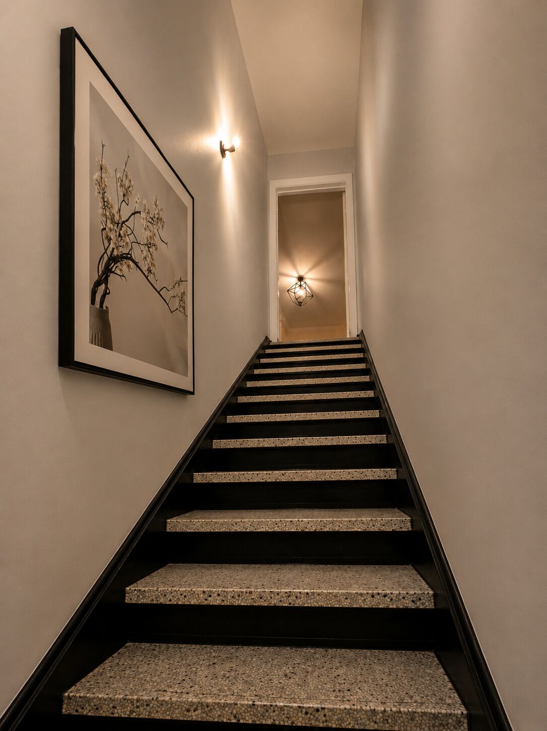 A staircase with a framed artwork on the wall, a light fixture above the door, and a textured floor.
