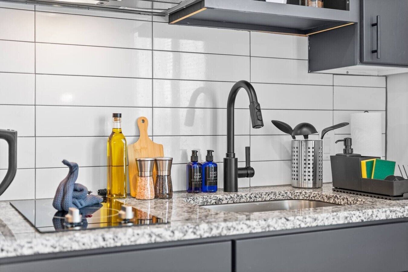 A kitchen with a modern design, featuring a stainless steel sink, a black faucet, and various kitchen utensils and bottles.