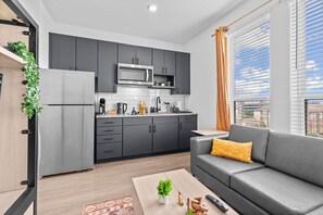 A modern kitchen with grey cabinets, a microwave, and a coffee machine. There is a grey sofa with a yellow pillow and a small table with a plant and remote control. - Sleek DT Studio | Rooftop Pool & Gym Access (Houston)