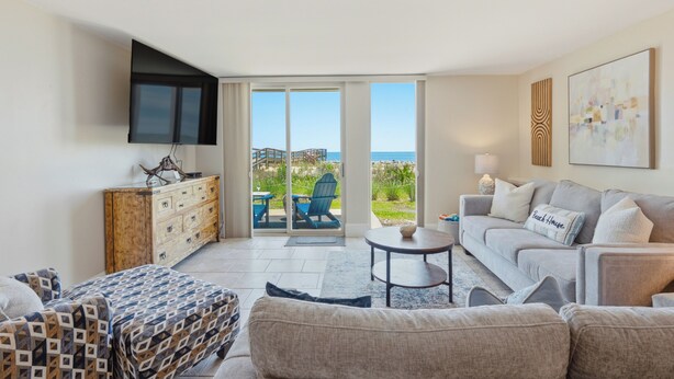A living room with a sofa, a coffee table, a TV mounted on the wall, and a view of the ocean through large sliding doors.