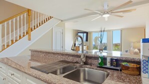A kitchen with granite countertops, stainless steel sink, wooden staircase, and a ceiling fan.