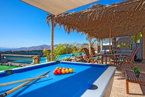 Ein Billardtisch mit Billardkugeln und Spielstöcken, ein Strohdach, Sitzgelegenheiten im Freien und Blick auf einen Pool und Berge.