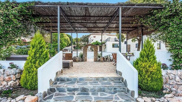 En stentrappa leder upp till en täckt uteplats med en pergola, omgiven av grönska och vita byggnader.