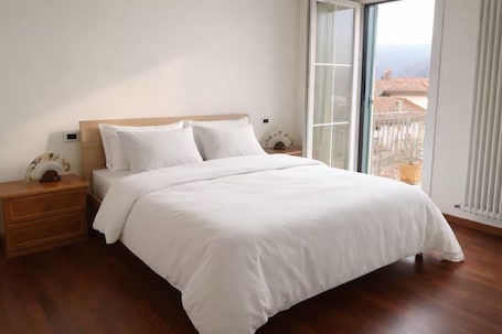 A bedroom with a wooden headboard, white bedding, a wooden nightstand, and a view of buildings through a large window.. Apartment with mountain view